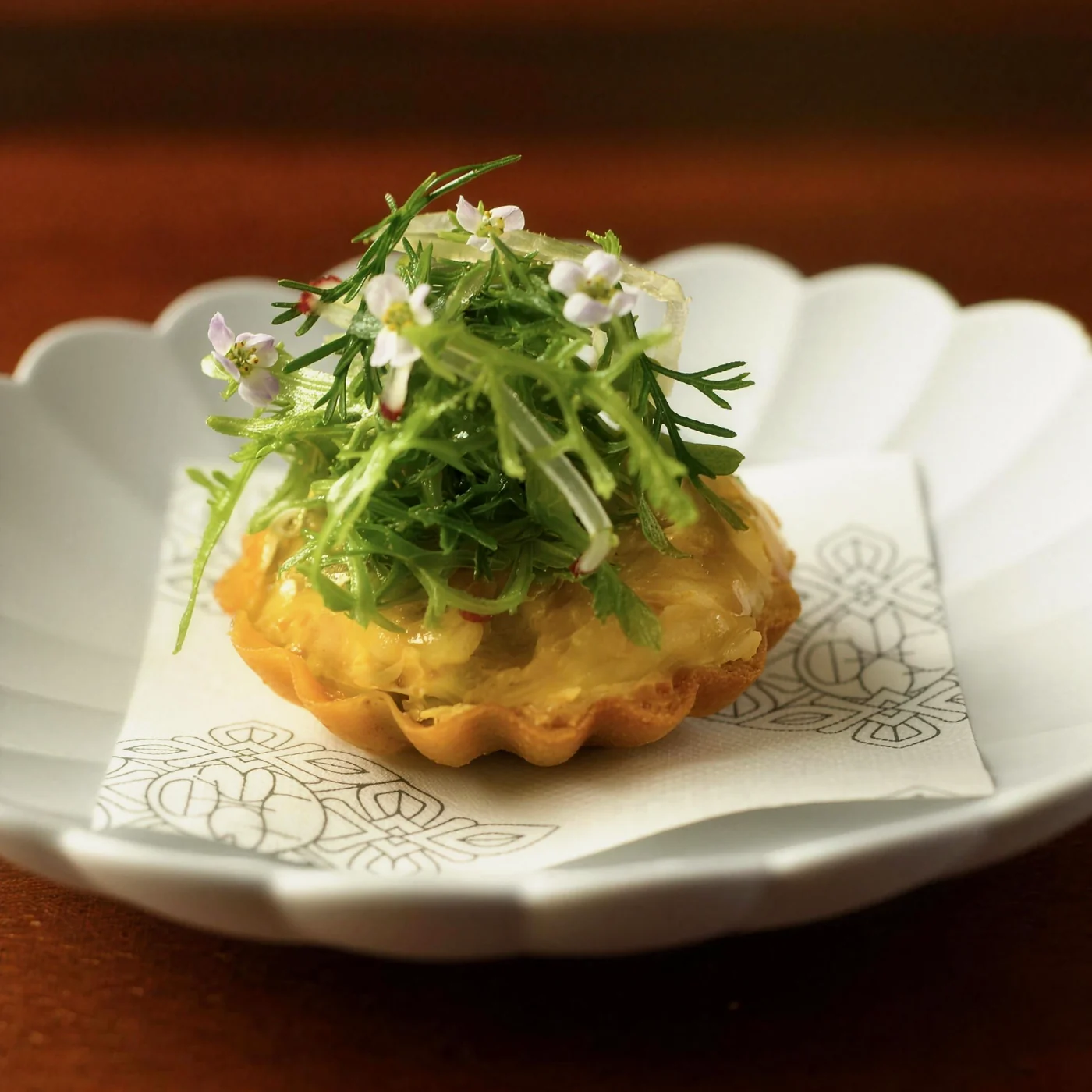 Golden tartlet crowned with fresh herbs and flowers on a scalloped white plate