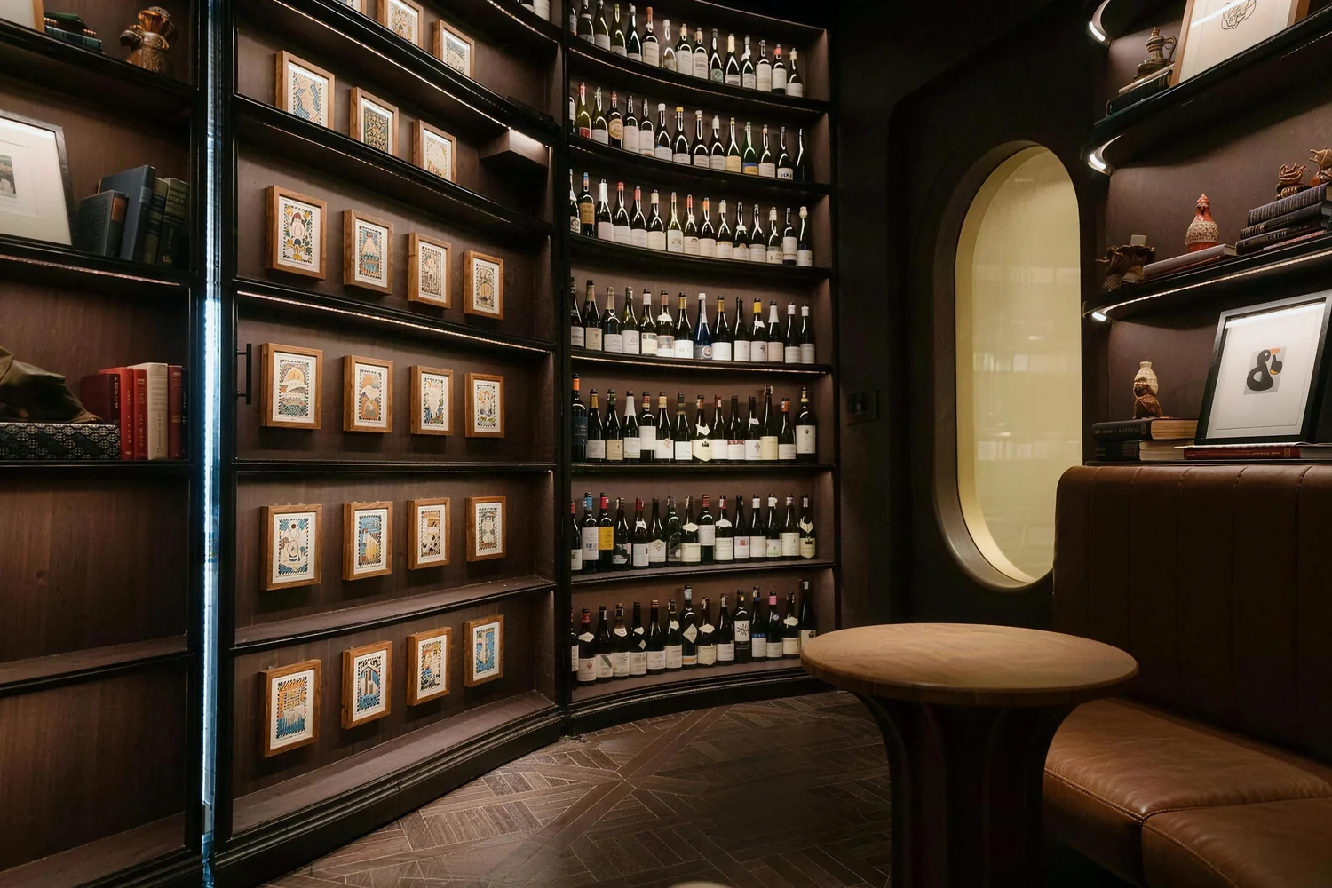 Curved dark-wood wine room with bottle-lined shelves, framed illustrations and a leather banquette