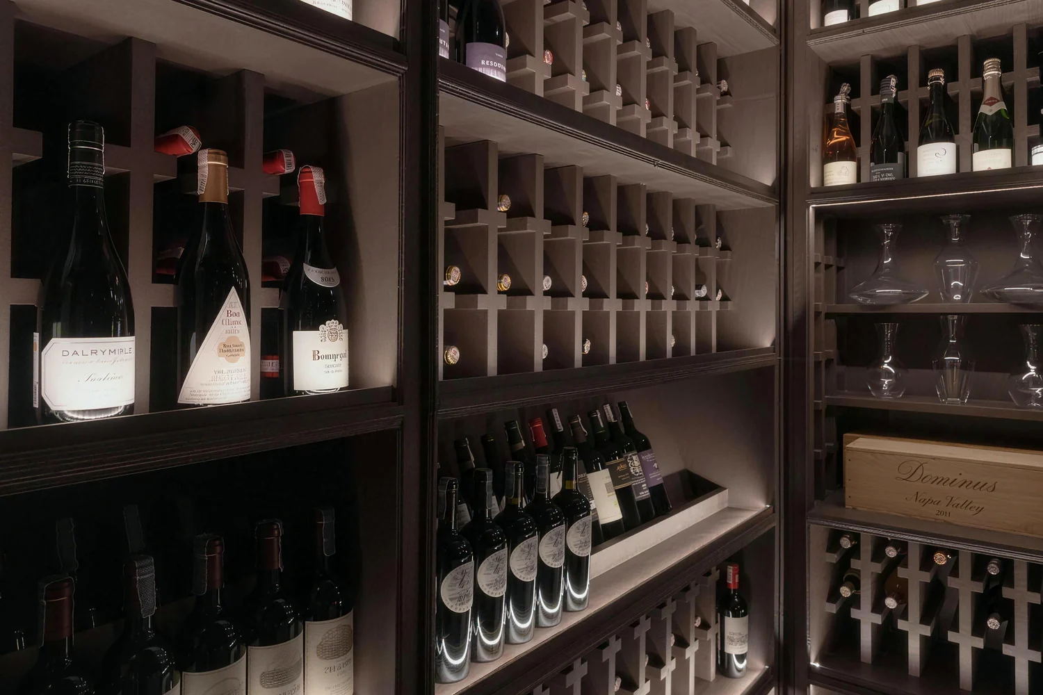 Backlit cellar racks filled with red and sparkling wine bottles, crystal decanters and a wooden case