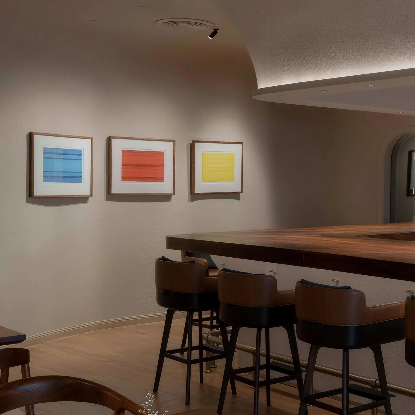 Curved dining room wall with three framed abstract prints above a walnut bar and leather stools
