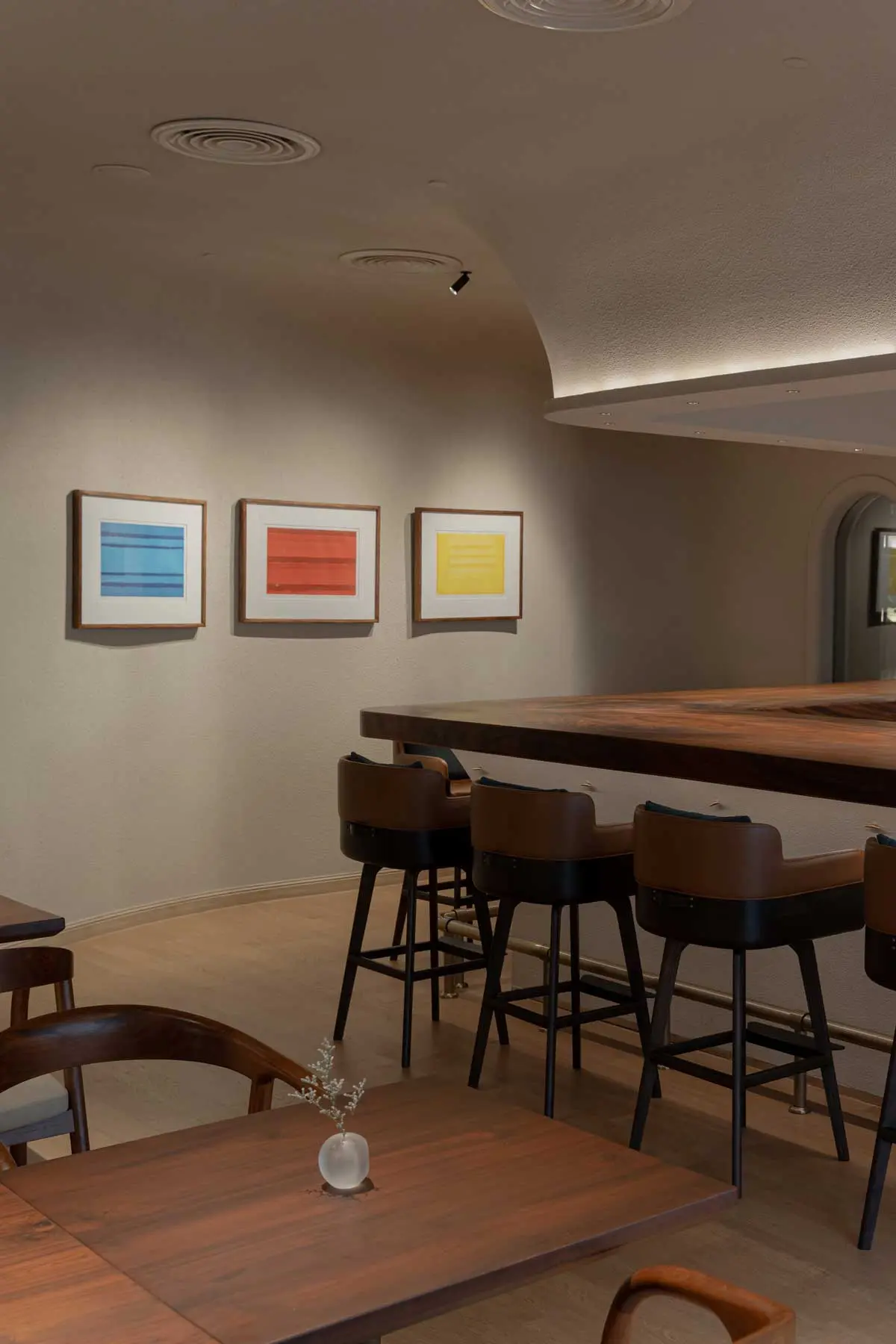 Curved dining room wall with three framed abstract prints above a walnut bar and leather stools