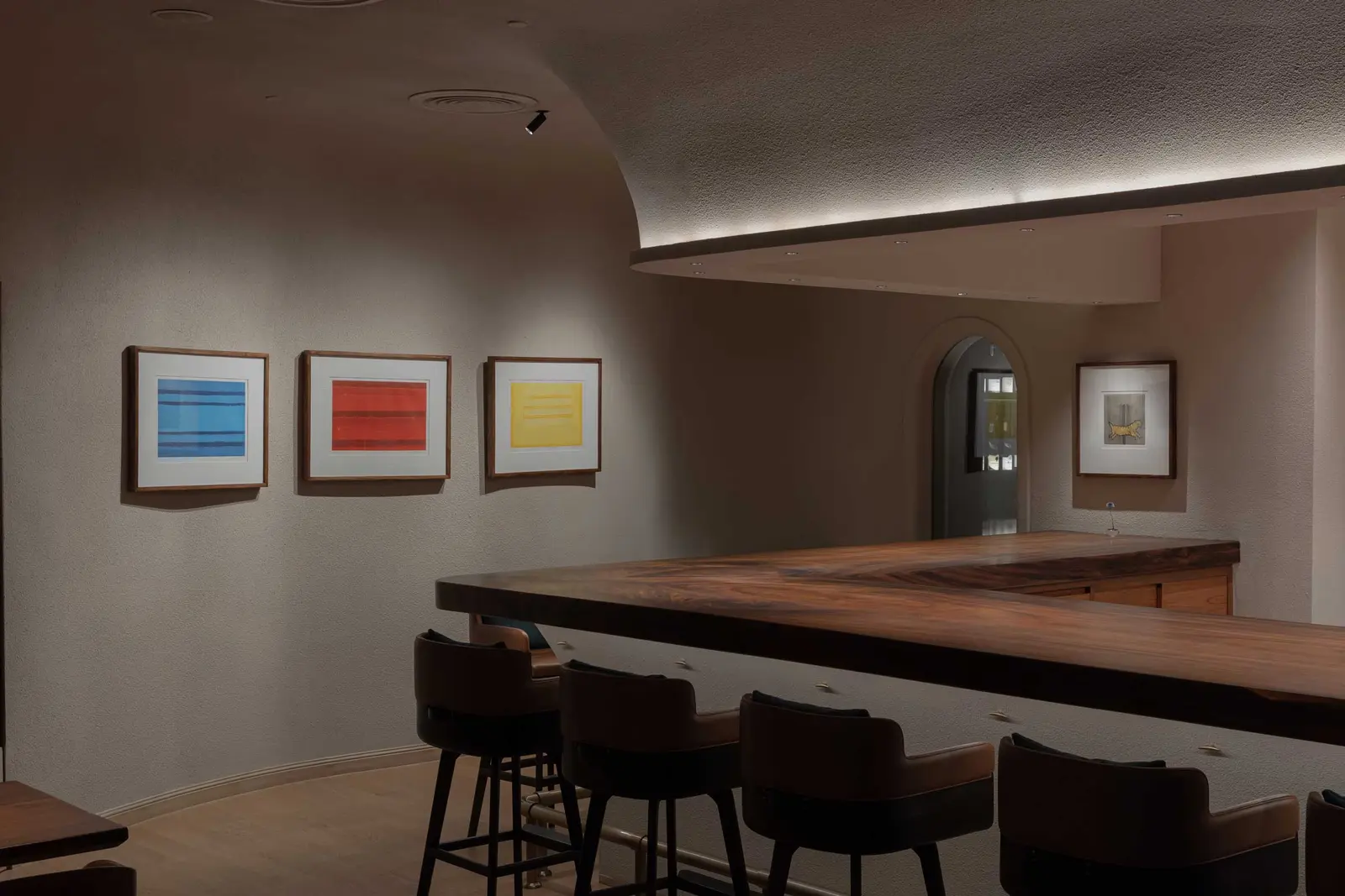 Walnut bar counter with leather stools below three framed blue, red and yellow prints